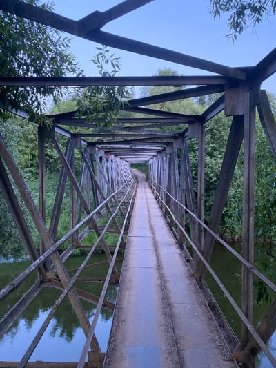 This is a vertical, eye-level, medium shot of a metal truss bridge spanning a body of water. The bridge is constructed of weathered steel beams and girders, forming a long, narrow walkway with a railing on either side. The walkway is made of metal plates, and it appears to be a pedestrian bridge, as there are no signs of vehicular traffic. The bridge is surrounded by lush green trees and foliage on both sides, with the water of a river or canal reflecting the trees and the sky. The sky is a pale blue with a few wispy clouds, suggesting it may be early morning or late afternoon. The bridge is in Yuryatino, Russia, and it is the Yuryatino footbridge. There are no people visible in the image, and no actions are taking place. The scene is one of natural beauty and rustic infrastructure.