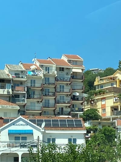 The image shows a multi-story apartment building with red tiled roofs in Meljine, Montenegro. The building is packed tightly with other similar structures, suggesting a dense urban environment. Several balconies are visible, some with chairs and tables, and laundry is hanging on one. Solar panels are installed on the roof of one section of the building. A sign on the front of this section reads "MAGNUM APARTMANI". The sky is clear blue, and lush green trees are visible in the foreground and background. No people are clearly visible in the image.