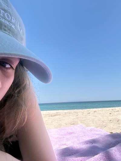 A low-angle, tilted close-up shot shows a person partially in frame, wearing a light green baseball cap with the word "TOPINO" embroidered on it, with "GENOVA" below. Their eye is visible, and their light brown hair is tousled. The person's arm is visible, resting or lying down. The background features a blue sky, a strip of blue-green water, and a beige sandy beach with a purple towel.
Based on the information visible on the baseball cap, the location is not within the city of Ολυμπιάδα, Greece. The cap says "Topino Genova", which refers to Genova, Italy. However, there is no information to confirm that the person in the image is located there.