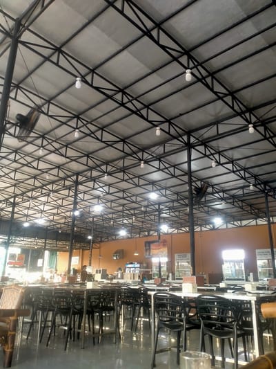The image shows the interior of a spacious, open-air restaurant or food court with a high, exposed metal roof structure. The roof is supported by black metal beams and trusses, with a light-colored, possibly insulated, sheeting. Multiple white, dome-shaped lights are suspended from the ceiling, and two large industrial fans are mounted on dark poles, angled downwards.

The main area of the restaurant is filled with rows of tables and chairs. The tables have white tops and silver metal legs. The chairs are black plastic, with a simple, modern design. Some of the tables are set with white placemats and small, white cube-shaped dispensers, possibly for napkins or sugar. In the foreground, a bamboo furniture piece is partially visible on the left, and a clear glass is on a table on the right.

The floor is a dark, reflective material, possibly polished concrete or tiles, mirroring the lights and furniture. In the background, there are advertisements and signage on the walls, some featuring food items like burgers and steak, suggesting it is a dining establishment. There are also glass display cases or counters, and what appear to be service areas or kitchens. A few people are visible in the background, indistinctly, either working or dining. The overall impression is of a well-lit, functional, and moderately busy casual dining space. The presence of advertising for "Azzam Home Eat" and "Mdm Burger" indicates this is likely a food establishment in the Medan Estate area of Indonesia.