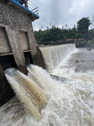 The image captures a dam spillway with water cascading over the edge and through two large openings in the stone structure. The water appears turbid and is generating significant foam and spray at the base of the dam. The dam itself is constructed of rough-hewn stones and concrete, with a blue railing visible at the top. Behind the dam, lush green vegetation, including palm trees, covers the landscape, suggesting a tropical environment. Several small buildings with thatched roofs are nestled amongst the trees. A bridge is visible in the distance, partially obscured by the foliage. The sky is overcast, indicating a cloudy day. This scene is consistent with the Tukad Unda dam in Klungkung, Bali, Indonesia, which is known for its tiered spillways and surrounding natural beauty. No people are visible in the image.