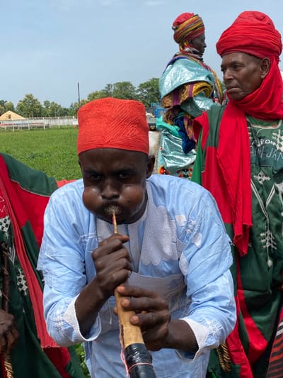 In Kafanchan A, Nigeria, a man in the foreground, wearing a red cap and light blue robe, plays a woodwind instrument. His cheeks are puffed out. Behind him are two more men in traditional attire, one with a red turban and the other with a multicolored headdress. All three are standing on a patterned cloth on a grassy field near a fence. In the background, there are trees and a light blue sky.