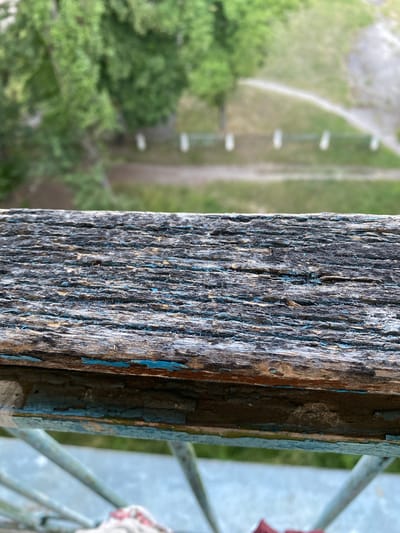 The image is a close-up shot of weathered, peeling blue and grey paint on a wooden beam. The wood is rough and textured, with deep grooves and cracks. The paint is flaking off in many places, revealing the wood underneath. In the background, out of focus, is a blurred green landscape with some linear structures that appear to be part of a park or outdoor recreational area. To the right of the beam, a blurred light grey metal railing is visible, and a small section of a white and red rope or fabric is also discernible. The lighting suggests it is daytime, and the overall impression is one of age and disrepair, with a hint of nature in the background. The location context is Shostka, Ukraine. No people, text, or specific activity are depicted.