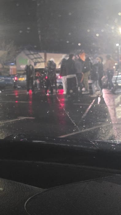 The image depicts a nighttime scene within a wet, paved car park or industrial area in Newry, United Kingdom. The view is from inside a vehicle, looking through a rain-streaked windshield. Approximately eight to ten individuals are congregated in the mid-ground, standing on the reflective asphalt. Most figures are clad in dark, indistinct clothing; one person on the far left wears a light-colored hooded garment. To the left of the group, a dark-colored vehicle is parked, displaying illuminated red taillights and a yellow license plate. Distant, indistinct buildings, one with a green-hued light, are visible in the background under artificial illumination, which also creates glare and reflections on the wet ground. The foreground displays the dark dashboard and the base of the windshield of the observation vehicle. No specific actions or interactions between the individuals are clearly discernible.