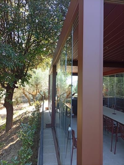 The image depicts an outdoor dining area with a covered patio and glass walls, situated in the city of Siena, Italy. The patio features a long table with multiple chairs, suggesting a restaurant or event space. The surrounding environment includes trees and foliage, indicating a natural or garden setting. The architecture suggests a modern design with clean lines and transparent elements.