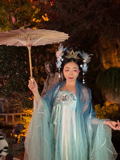 In the city of Unknown, China, a young woman in traditional Chinese attire stands holding an umbrella. She wears a blue and white embroidered dress and a delicate headpiece adorned with flowers. The background is a dimly lit garden with lush greenery and illuminated trees, creating a mystical ambiance. A statue of a person is visible behind her, adding to the historical feel of the scene.