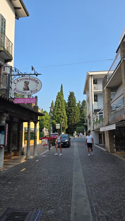 The image depicts a street scene in Sirmione, Italy, likely near the lakefront given the sign "GIARDINO VISTA LAGO" (Garden Lake View). The cobblestone street is lined with buildings on both sides. On the left, a building with a facade of brick and stucco houses a restaurant with a sign reading "L'Arcimboldo Ristorante con Pizza". The restaurant has a covered outdoor seating area supported by pillars. On the right, a multi-story building, identified by a sign as "HOTEL LUNA", has balconies with white railings.

In the mid-ground of the street, two people are walking. A woman in a white tank top and denim shorts walks towards the foreground, and a man in a white polo shirt and dark shorts walks away from the foreground. A dark-colored car is parked on the right side of the street near the woman. Further down the street, other pedestrians can be seen, and a row of tall, slender cypress trees stands behind the buildings on the left. The sky is clear and blue. The overall impression is a sunny day in a European town.