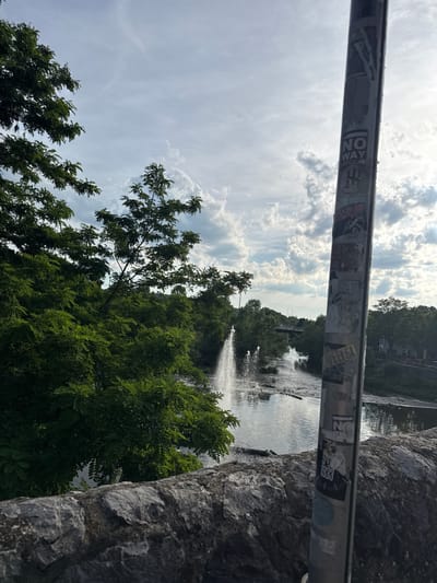 The image shows a vertical view of a canal or river, with a prominent metal pole in the foreground. This pole is adorned with numerous stickers, some of which are partially visible and contain text. One sticker clearly reads "NO WAY". The water has a fountain spraying in the middle distance, creating a white mist. Lush green trees line the banks of the waterway, with more foliage appearing in the foreground, obscuring parts of the scene. A stone wall is visible on the right side of the frame. The sky is filled with dramatic, cloud formations, suggesting a dynamic weather pattern. The overall impression is one of an outdoor scene, possibly in a park or along a riverfront, with the sun partially obscured by clouds, creating a bright but not overly sunny atmosphere. The presence of stickers on the pole suggests an urban or public space where such decorations are common. The text on the stickers adds a touch of cultural commentary or personal expression to the scene. The image's orientation is vertical, which emphasizes the height of the pole and the expanse of the sky. The presence of a fountain indicates a likely public amenity. The overall mood is serene, with the natural elements of trees and water contrasting with the man-made elements of the pole and fountain. There are no people visible in the frame.