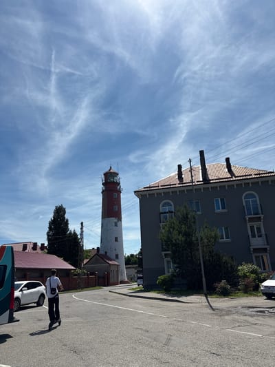 A tall, red and white lighthouse stands prominently against a bright blue sky with scattered white clouds. To the right of the lighthouse is a multi-story building with a gray facade, a red tiled roof, and arched windows. In the foreground, a person is riding an electric scooter on a paved road. Several cars and other buildings with red roofs are visible in the background. The scene appears to be in a coastal or harbor area, consistent with the location of Baltiysk, Russia, a port city known for its lighthouse. The lighting suggests it is daytime with clear weather.