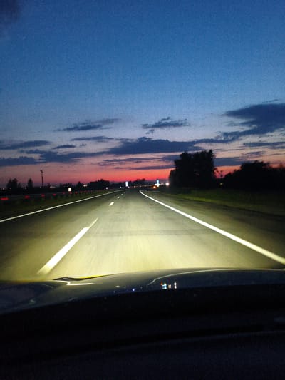 This is a photograph taken from inside a vehicle at dusk, driving on a highway in Gramoteino, Russia. The perspective is from the driver's seat, looking forward along the road. The sky is a gradient of deep blue at the top, transitioning to shades of pink and orange near the horizon, indicating the sunset or twilight. Wispy clouds are scattered across the sky.

The highway is well-lit by the vehicle's headlights, which illuminate the asphalt and the bright white lane markings. The road appears to be a multi-lane highway, with a clear divider. On the sides of the road, there are trees and vegetation, silhouetted against the colorful sky. There are also guardrails with reflective markers visible along the side. In the distance, some lights suggest the presence of signs or structures, possibly related to the highway or a nearby town. The overall mood is peaceful and solitary, evoking the feeling of a late-evening journey. There is no visible text in the image.