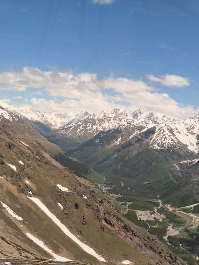The image captures a breathtaking panoramic view of a mountain valley, likely in or near Terskol, Russia. The foreground is dominated by a steep, rocky mountainside with patches of snow scattered across its brown and green terrain. Several parallel wires, possibly from a ski lift or cable car, traverse this slope diagonally.

Further into the scene, the valley opens up, revealing a lush green landscape dotted with trees. A winding river or streambed carves through the valley floor, its path marked by lighter colored sand and gravel. In the distance, nestled within the valley, a small town or settlement can be observed with buildings and infrastructure.

The background is filled with majestic, snow-capped mountain peaks that stretch across the horizon. Their rugged slopes and white summits create a dramatic contrast with the vibrant green of the valley below. The sky above is a clear blue, adorned with scattered white clouds, some of which appear wispy and elongated. The overall impression is one of a vast, natural landscape, highlighting the grandeur of the Caucasus Mountains.