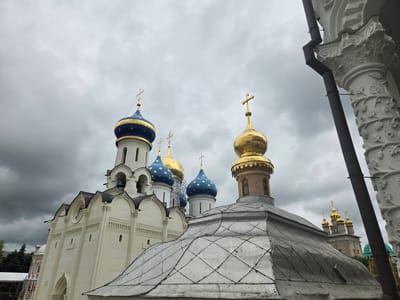This media file captures a close-up, low-angle view of the Trinity Lavra of St. Sergius in Sergiyev Posad, Russia, under a cloudy sky. The primary subjects are the distinct domes and architectural features of several churches within the monastery complex.

The image prominently displays several domes: a large, white church with multiple arched openings and bell towers, topped by a blue dome with gold stars and a gold cross; adjacent to it, another blue-domed structure with gold stars; and a bright gold dome with a gold cross. To the right, a portion of a richly decorated white column with carved floral motifs is visible, alongside a dark pipe running vertically. In the foreground, a large, weathered metal roof with a diamond-patterned texture dominates the lower part of the frame. Further in the background, several smaller golden domes of another church are visible, hinting at the vastness of the monastic site.

The weather appears overcast and possibly stormy, with heavy grey clouds filling the sky. The lighting is diffused and subdued. There is no apparent activity or event taking place, as no people are visible. The overall mood is solemn and perhaps a bit dramatic due to the cloudy sky. The style of the architecture, particularly the onion domes and ornate stonework, clearly indicates a Russian Orthodox setting. No visible text is present in the image.