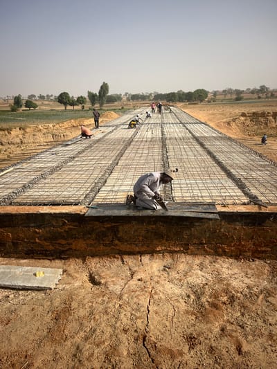 Several workers are assembling rebar grids, likely for a concrete pour, on a large outdoor construction site. A man in a light-colored outfit and hat sits on the edge, working on the steel reinforcement with tools in hand. Long rows of the grid extend into the distance, with a few other workers positioned along the length. The construction area is bordered by earthen walls and the surrounding landscape includes agricultural fields.