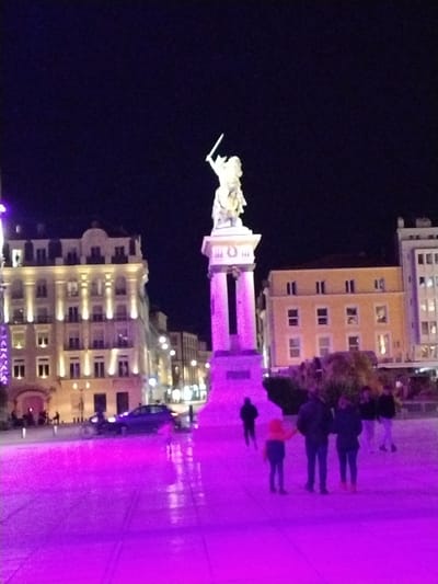 The image displays a nighttime urban scene in Place de Jaude, Clermont-Ferrand, France. The central feature is the equestrian statue of Vercingetorix, depicting him with a sword raised, positioned on a tall, multi-part stone pedestal. The lower portion of the pedestal is illuminated by intense purple light, which also bathes the paved square in the foreground and midground, reflecting off a visibly wet surface.
To the left of the statue, a multi-story building with classical architecture and multiple lit windows is illuminated by warm white external lighting. To the right, a plainer multi-story building also exhibits illuminated windows. A dark-colored sedan is visible in the midground, positioned left of the statue's base.
Approximately seven individuals are present. In the right midground, a group of three individuals—two adults and one child in a bright orange top—stand on the purple-lit pavement. A single, silhouetted individual walks away from the viewer in the left midground. Additional indistinct figures are visible further in the background. The sky is dark and clear, indicating nighttime conditions.
