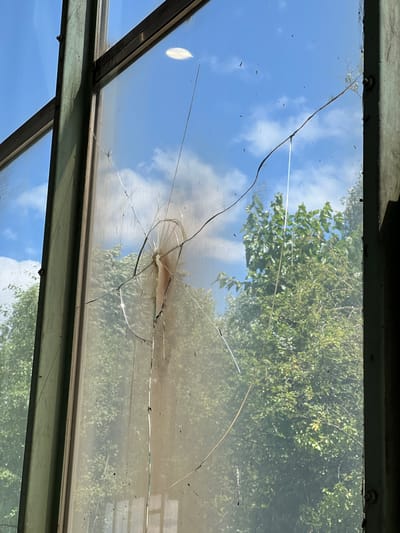 The image shows a close-up view of a broken window pane, likely from an abandoned or industrial building in Lanžhot, Czechia. The window frame is a weathered, dark green metal. The glass is cracked in multiple places, with a spiderweb pattern radiating from a central impact point. The cracks are sharp and jagged. Dirty streaks and small dark particles are visible on the glass surface.
Through the broken window, a clear blue sky with scattered white clouds is visible. Lush green trees, with dense foliage, form the background. The lighting suggests it is daytime, with sunlight filtering through the leaves and reflecting off the glass. There are no people or animals visible in the image. The scene conveys a sense of decay and neglect.