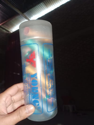 The image shows a person's hand holding a translucent plastic bottle filled with small, colorful objects. The bottle has text and a logo printed on it. The logo appears to be a red stylized 'Y' shape above the word "YOUNG" in blue letters. The text on the bottle is in Indonesian and includes "Kain Lap Serbaguna" (Multipurpose Cleaning Cloth), "Cocok untuk: Komputer, Mainan, Sepeda, Motor, Mobil, etc." (Suitable for: Computers, Toys, Bicycles, Motorcycles, Cars, etc.), and dimensions "12 x 42 x 0.2 CM". The background is dark with some light illuminating a corrugated metal ceiling. The scene is likely indoors, possibly a storage area or a shop, in Cotseutui, Indonesia.