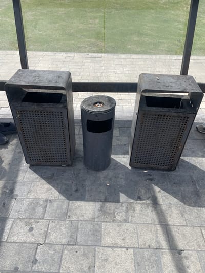 The image shows three outdoor trash receptacles on a paved area. Two of the receptacles are rectangular with open slots on the front and a perforated metal top. The third, a cylindrical bin, is situated between the two rectangular ones and has an open slot on its side. The ground is paved with gray square tiles. To the left of the paved area is a green grassy field. A thin black metal railing runs horizontally across the top left of the frame, separating the grassy area from the paved area. The scene is lit by bright sunlight, casting shadows on the ground, indicating it is daytime. There is no visible text in the image. The overall impression is one of an outdoor public space, possibly a park or a recreational area, with an emphasis on cleanliness and waste disposal.