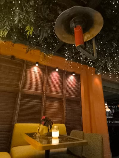 This image depicts a warm and inviting interior or semi-outdoor seating area, characteristic of a restaurant, bar, or cafe, likely in a location such as Palaio Faliro, Greece. The scene is set during the evening or night, illuminated by various artificial light sources.

The ceiling is adorned with lush artificial greenery, densely woven with numerous small, twinkling string lights that create a festive and magical ambiance. A large, sombrero-shaped outdoor heater with a glowing red heating element hangs prominently from the ceiling, suggesting the space might be open-air or requires supplemental warmth.

In the foreground, a small square table with a dark, reflective surface is positioned between two upholstered seating options: a mustard-yellow booth in the immediate foreground and another partially visible behind the table. A small potted plant with reddish-brown leaves and a delicate candle flickering in a glass holder grace the tabletop, adding a cozy detail. Part of a textured green armchair is visible to the right of the table.

The walls feature a mix of materials and colors. The background is dominated by warm brown wooden panels, possibly decorative shutters or slatted screens, which are softly illuminated by three recessed spotlights casting reddish hues. An upright pillar and a section of the wall on the right are painted in a vibrant orange or terracotta shade, contributing to the Mediterranean aesthetic. Blurred elements in the deeper background suggest other parts of the establishment or an outdoor view. No people are visible, emphasizing the serene and prepared atmosphere. There is no visible text in the image.