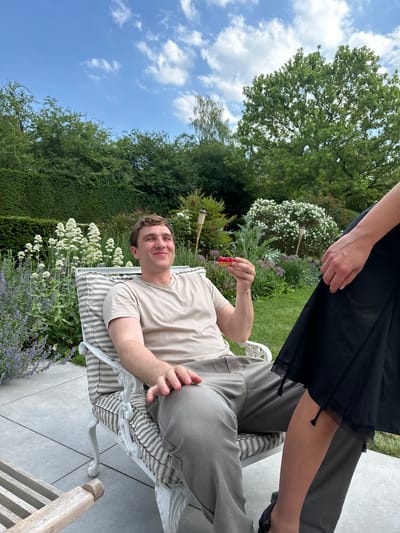 The image shows a person lying on a striped lounge chair in a garden. The person is fair-skinned, wearing a beige shirt and gray pants, holding a small red dessert or fruit in their right hand. Another person partially visible at the top of the frame is wearing a black dress. The garden features a manicured hedge, white and lavender flowers, and various plants. The sky is blue with scattered white clouds. While it is not possible to definitively ascertain the location as being in Cambridge, UK, the garden style and architecture are consistent with residential areas in that city.