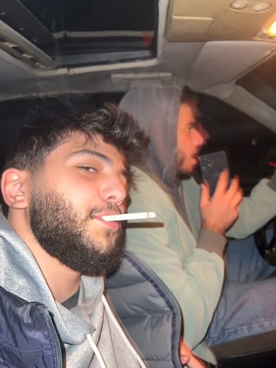 Inside a vehicle, two bearded men are visible. The lower man has a cigarette in his mouth and is facing the camera. The man behind him is holding a phone and looking down at the screen. He's wearing jeans and a green jacket. The interior is a pale color with an overhead light. The image is slightly rotated.