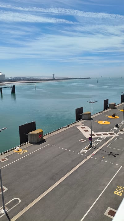 The image depicts a waterfront scene with a paved area on the right, likely a port or industrial waterfront. The paved surface has various markings, including white lines indicating lanes and parking spots, as well as yellow stenciled numbers and symbols. Notably, the number "B06" is visible twice, and other yellow circular markings are present. Several concrete blocks with yellow tops and red circular markers are strategically placed along the edge of the paved area, possibly as bollards or safety barriers.

To the left of the paved area is a vast expanse of calm, turquoise water. In the distance, a hazy shoreline is visible with some structures, including what appears to be a tower or industrial building. The sky above is a brilliant blue, streaked with thin, wispy white clouds, suggesting fair weather and possibly mid-morning or afternoon. The overall impression is one of a working port or industrial waterfront under clear skies. There are no people prominently visible in the image, nor is there any clear activity or event taking place. The scene is static and focuses on the infrastructure and the natural elements.