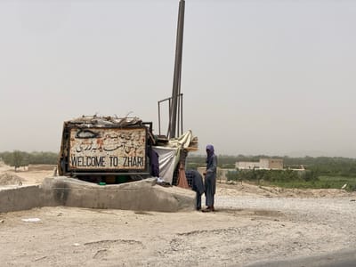 This image depicts a dusty, sunlit outdoor scene, likely a roadside or entrance point to a settlement. The sky is hazy and light-colored, indicating a dry environment.
**Central Subject:** A prominent, weathered metal sign structure, likely a welcome sign, is positioned on a concrete base on the left side of the frame. The sign displays text in both Pashto and English, with the English portion clearly reading "WELCOME TO ZHARI". The structure appears makeshift, with various metal components and debris on top. Behind this sign, a tall, dark metal pole stands upright.
**People:** Two individuals, presumably males, are visible to the right of the sign. They are dressed in long, light-colored traditional garments, possibly shalwar kameez. The individual on the left stands upright, facing right, with a dark head covering. The individual on the right is bent forward, appearing to be interacting with something low to the ground or within the sign's structure.
**Environment:** The immediate foreground consists of unpaved, light-colored ground, indicating a dirt or gravel road. In the mid-ground, a single-story building with a flat roof is visible amidst some sparse vegetation. Further in the background, a band of denser green vegetation or trees stretches horizontally, suggesting cultivated land or natural greenery, leading to a hazy horizon with indistinct landforms.
**Location:** Based on the prominent sign, the scene is at an entrance or boundary to Zhari, Afghanistan. Zhari is a district located in Kandahar Province. The image does not show details identifying the specific location as within the city of Dand, Afghanistan, but rather indicates Zhari.