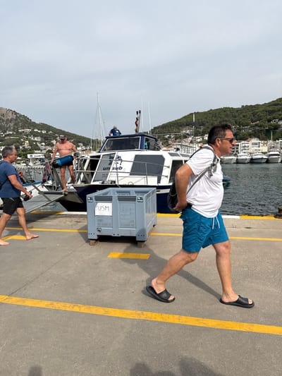 The image presents a daytime scene at a marina in Torroella de Montgrí, Spain. A light-skinned man with short brown hair, wearing a white t-shirt, blue swim shorts, and black slide sandals, walks left-to-right across a concrete pier in the foreground. He carries a dark shoulder bag over his left shoulder. To his left, a large grey plastic crate marked "USM" in white stands on wooden blocks on the pier.
Behind the crate, a white and dark blue motorboat or catamaran is docked. The boat's hull displays "un sub" and "2-7" text, along with a telephone number and website address. On the stern platform of this boat, a shirtless man wearing blue swim shorts and a baseball cap holds a dark blue cylindrical object. Another man, wearing a dark blue shirt, is partially visible standing on the boat's cabin roof. A third man, light-skinned with short dark hair, wearing a blue t-shirt and black shorts, stands barefoot on the pier to the left of the boat, appearing to interact with the vessel or an unseen object.
The background features a marina with numerous other boats docked in calm water. Beyond the marina, green, tree-covered hills rise, dotted with white and light-colored buildings. The sky is partly cloudy, and sunlight illuminates the scene, casting shadows on the pier. The concrete pier surface features two horizontal yellow painted lines.