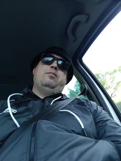 A man wearing sunglasses and a black bucket hat is in the driver's seat of a car, looking up towards the camera. He is wearing a black jacket with white drawstrings and a seatbelt is across his chest. The interior of the car is visible, including the handle above the door and the door frame. Outside the car window, trees and sky are visible, suggesting an outdoor, potentially rural or suburban setting. The reflection in the sunglasses shows a road with trees and a clear sky, which could indicate the vehicle is in motion or parked on a road. The image was taken on the street of Brandýs nad Labem-Stará Boleslav, Czechia.