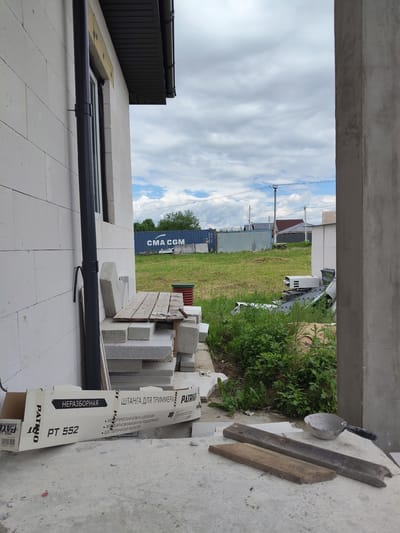 In Skurygino, Russia, an exterior shot captures a construction site on a cloudy day. A pale brick building, possibly under construction, dominates the left side of the frame, featuring a dark downspout running vertically. In the foreground, a cardboard box with Russian text, including "ПАТРИОТ" and "PT 552," lies on a concrete surface. To the right, a concrete pillar marks the edge of a building or structure. Beyond the immediate construction area, a grassy field is visible, dotted with green vegetation. In the middle ground, a blue shipping container with "CMA CGM" printed on it stands alongside a light gray fence. Further in the distance, houses with dark roofs and utility poles with power lines suggest a residential or semi-rural area. Several stacks of concrete blocks and wooden planks are also present on the site, indicating ongoing construction work.