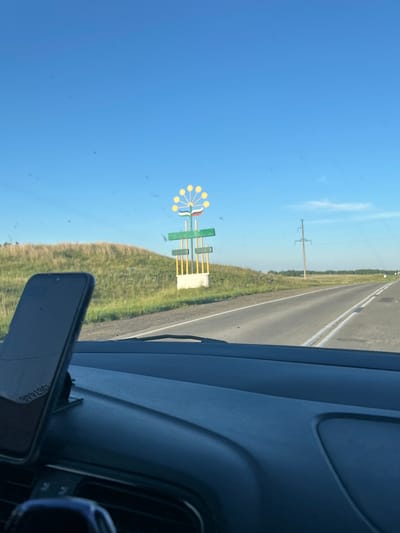 A daytime, eye-level, over-the-shoulder shot captures a rural roadside scene through a car windshield. In the foreground, the dark dashboard of a vehicle is visible, with a smartphone mounted on the right side. Beyond the dashboard, a two-lane asphalt road stretches into the distance, bordered by green fields and a gently sloping grassy hill on the left. A tall, decorative signpost resembling a stylized flower with yellow petals stands prominently on the hill. Further down the road, a utility pole is visible on the right. The sky is a clear, bright blue. The image is believed to be taken in or near Ishmurzino, Russia, based on the sign's likely representation of a local landmark or symbol.