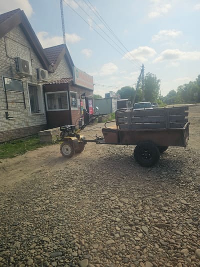 A yellow motor cultivator with a trailer is parked on a gravel road in front of a small brick building with a wooden addition. The building has signs that read "СВЕЖИЙ" and "МАГАЗИН САМООБСЛУЖИВАНИЯ". There are two air conditioning units mounted on the brick wall. The trailer is filled with wooden planks. In the background, there are trees and a white pickup truck. The sky is blue with some white clouds. The overall scene suggests a rural or semi-rural setting, possibly a small shop or convenience store.