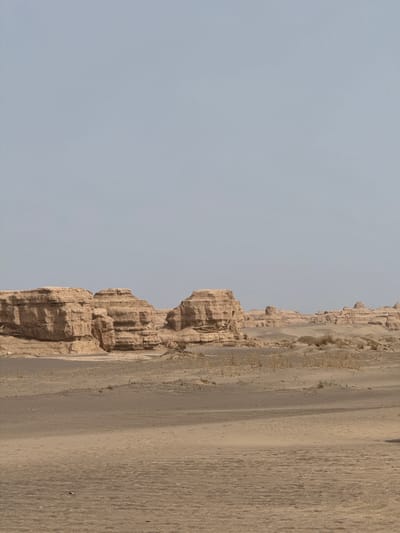 The image shows a geological formation in Dunhuang City, China. The formation is composed of layered rock with a heavily eroded surface, abutting a sandy slope. The sky is overcast and uniformly grey. There are no signs of human or animal activity in the frame.
