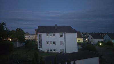 The image captures a twilight scene in a residential area, likely within the city of Zweibrücken, Germany, given the architectural style of the houses and the overall atmosphere. The sky is a deep, cloudy blue, indicating the late evening or early morning hours. In the foreground, a dark-tiled roof of a building is visible, suggesting this is an elevated viewpoint.

The central focus is a three-story, white-painted apartment building with a dark grey, pitched roof and a prominent grey chimney. This building features a grid of small, square windows on the left side and rows of rectangular windows on its facade. Some windows have blinds.

To the right of the main building, a street is partially visible, illuminated by a yellow streetlamp. Several other residential houses are scattered across the landscape, with pitched roofs of varying colors, including white, yellow, and blue. These houses appear to be on a gentle slope, leading up to a more densely populated area in the distance, where more rooftops and lights can be seen.

In the mid-ground, there are lush green trees and bushes, some casting dark shadows. A smaller, light-colored building or garage is situated to the left, with scaffolding visible behind it, suggesting ongoing construction or renovation. A white van is parked near this structure. The overall environment is serene and quiet, characteristic of a suburban or semi-urban setting during the transition from day to night. There are no people visible in the image.