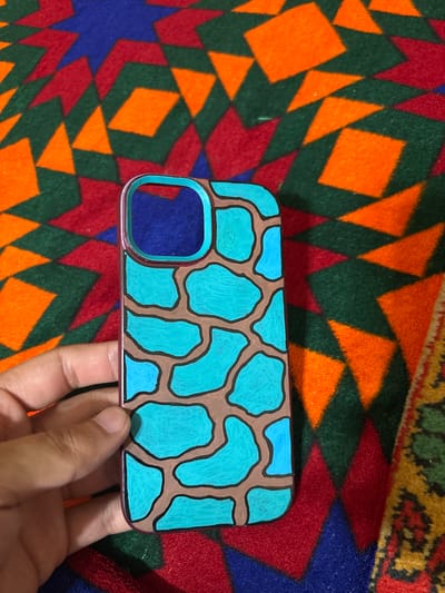 A hand holds a phone case with a light blue, irregular pattern outlined in dark brown, resembling giraffe spots. The phone case itself is a light pinkish-purple color and has a cutout for the camera lens. The background is a colorful, geometric textile with diamond and triangular patterns in shades of orange, green, red, and blue. The lighting suggests it's daytime, likely indoors given the close-up view of the object and hand. The overall mood is casual and focused on the custom phone case. No visible text is present. The provided location context of Nukus, Uzbekistan, does not offer specific visual cues within the image itself.