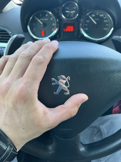 The image displays the interior of a vehicle, specifically a Peugeot, from the driver's perspective in Maksimkovo, Russia. A light-skinned human hand, exhibiting a dark leather wristband with white stitching on the wrist, rests on the black, textured steering wheel. The silver Peugeot lion emblem is centrally located on the steering wheel's airbag cover, with the word "AIRBAG" embossed below it. Behind the steering wheel, the instrument cluster is visible, showing a tachometer with the needle near the idle position and a digital "S" indicator. A central digital display shows red numerals, potentially indicating time or an error code. The speedometer on the right indicates a near-zero vehicle speed. A paddle shifter is discernible on the left side, behind the steering wheel. A portion of a leg wearing light-colored trousers is visible below the steering wheel, indicating the presence of a driver. Natural light illuminates the cabin.