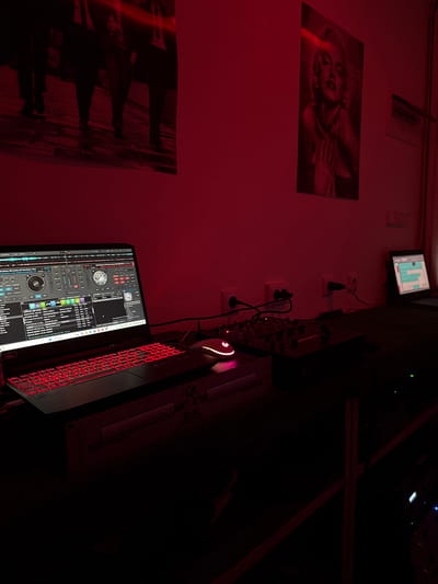 The image depicts a dimly lit room, possibly a DJ booth or a small studio, with a strong red ambient light. On a dark surface, a laptop with a red illuminated keyboard is open, displaying DJ software with waveforms and track lists. A wireless mouse with a glowing red scroll wheel rests to the right of the laptop. In front of the laptop, there is a DJ mixer with multiple knobs and faders. Further to the right, a second, smaller laptop or screen is visible, displaying a grid-like interface, and behind it, an electrical outlet with a plug. The wall behind the equipment is painted red and features two framed prints: one on the left showing the lower bodies of four men in suits walking, and another on the right, a portrait of Marilyn Monroe. Electrical outlets are visible on the wall, some with plugs inserted. The overall impression is of a space set up for music production or performance, with a retro or artistic aesthetic. There are no people present in the image.