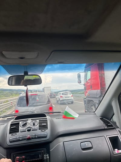 The image shows a view from inside a vehicle stuck in traffic on a highway. A red truck is visible to the right, and other cars are ahead on the road. A small Bulgarian flag is placed on the dashboard. The dashboard of the vehicle is visible in the foreground, with various controls and an audio system. The exterior view through the windshield reveals a cloudy sky and a scenic landscape with hills in the distance. The traffic appears to be moving slowly or is stopped, characteristic of congestion. The presence of the Bulgarian flag suggests the location might be within Bulgaria. Based on external information, the image was taken on the A1 highway near Vakarel, Bulgaria.