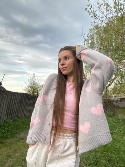 A young woman is the central subject of the image, posed outdoors against a backdrop of sky, trees, and vegetation. She is wearing a pink top, light-colored sweatpants, and a gray cardigan adorned with pink hearts. Her long, straight hair is loose around her shoulders. Her hand is raised near her head. The background includes trees, a cloudy sky, and what appears to be a corrugated roofing material in the lower part of the frame. The location is identified as Борщевка Малая, Russia.