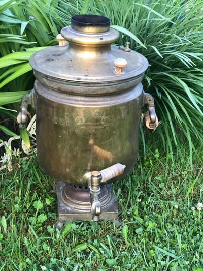 An antique, brass samovar stands on a square base in a grassy area, surrounded by lush green foliage. The samovar has a wide body with two wooden handles on the sides and a spigot near the bottom. It is topped with a lid and a chimney. The details on the samovar indicate it is an ornate piece, possibly with engravings. The background is filled with the dense green leaves of what appear to be daylilies or similar plants. The ground is covered in a mix of grass and small clover plants, with a few tiny white wildflowers visible. The lighting suggests it is daytime, with sunlight casting highlights on the metal surfaces of the samovar. The overall impression is of a historical artifact placed in a natural setting, possibly in a garden. The image does not contain any people or animals, nor any specific urban or rural landmarks that would definitively place it in Staroe Istomino, Russia, or any other specific city.