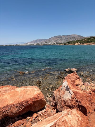 This media file depicts a coastal scene in the Vouliagmeni Municipal Unit, Greece. The image is oriented vertically, showing a view of the sea from a rocky shoreline. The foreground is dominated by large, reddish-orange rocks, some of which are partially submerged in the clear, turquoise water. The water itself transitions to a deeper blue further out. The seabed is visible through the shallow water, appearing rocky and pebbly.
In the mid-ground, a town is nestled along the base of a steep, verdant hill. The buildings are densely packed and appear to be white or light-colored. The hill rises behind the town, covered with sparse vegetation and rocky outcrops. The sky is a clear, bright blue, suggesting a sunny day. The sun appears to be high in the sky, casting sharp shadows on the rocks.
The scene captures a tranquil and picturesque coastal landscape. There are no people or specific objects of note, other than the natural elements of rock, water, and vegetation, and the man-made town. The overall impression is one of a warm, dry day in a Mediterranean coastal region. There is no visible text in the image.