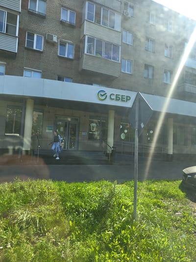 The image shows a Sberbank branch located in Zhukovsky, Russia. A woman wearing jeans and a dark top, carrying a large blue and white patterned bag, is walking out of the bank. She is on the steps leading out of the entrance, with her right foot forward. To the left of the entrance, a pillar supports the awning of the building. The bank's signage, featuring the Sberbank logo and text, is visible above the entrance. The building itself appears to be a multi-story residential building with cream-colored brickwork and multiple windows. In front of the building is a strip of green grass, and beyond that, a paved area, possibly a sidewalk or road. Sunlight is strongly illuminating the scene from the right, creating lens flare and reflections. A dark-colored car is partially visible on the right side of the image, parked on the paved area. A traffic sign on a metal pole stands in the foreground, slightly off-center.