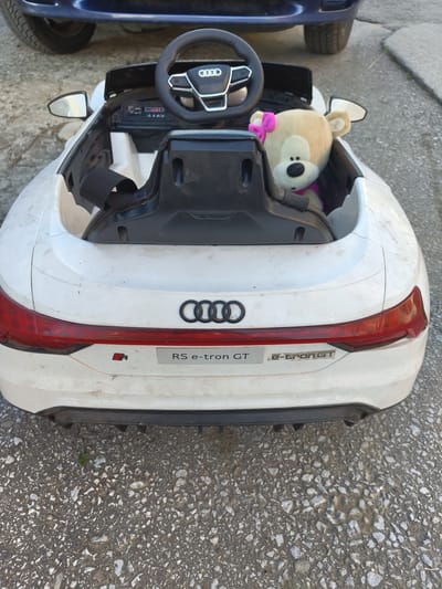 A white electric Audi RS e-tron GT toy car, with a teddy bear inside, is parked on a paved surface in Pletena, Bulgaria. The car is dirty and appears weathered. A portion of a dark blue vehicle is visible in the upper background. The teddy bear is light brown with a pink bow on its head and a purple scarf. The car's interior includes a black steering wheel with the Audi logo and a black seat with a strap. The rear of the car features the Audi rings logo, an "RS e-tron GT" badge, and red taillights. The ground is a mix of asphalt and small stones.