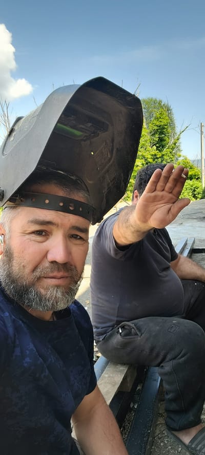 Two men are visible in this outdoor, daylight selfie. The man in the foreground, facing the camera, wears a dark blue t-shirt with a camouflage pattern and a welding mask on his head. He has a grey beard and appears to be looking towards the camera. The man in the background, partially obscured, wears a dark t-shirt and dark pants. He is seated and holding his right hand up, obscuring part of his face and the camera's view, possibly holding a cigarette. They are outdoors, with a blue sky and some clouds visible, along with green trees in the background. The ground appears to be gravel and dirt, and there are some metal and wooden objects around them. The location is specified as Milisi, Georgia.