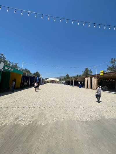 A wide-angle, eye-level outdoor shot depicts a temporary market or event area in Daranak, Armenia, under a clear, cloudless blue sky. The scene features a broad, light-colored gravel path spanning from the foreground to the distant background. Along both sides of the path are rows of semi-permanent structures or booths.

On the left, structures are primarily colored in green-teal and yellow, with a prominent "acba" logo visible on the green-teal section of the nearest booth. One individual, appearing male, is visible near the yellow section of this booth. Further along the left, another person is partially visible behind a dark structure. Overhead, multiple strands of string lights with numerous white, round bulbs are suspended between poles, forming an archway over the path. Pine trees are visible behind the structures on both sides.

On the right, structures are uniformly designed with light brown fabric partitions or walls, resembling shaded outdoor seating areas. A yellow sign bearing the text "YO HEALTH" is visible on the nearest structure. Several individuals are seated or standing within this "YO HEALTH" area.

In the mid-ground, a woman wearing a white top and dark skirt/shorts walks away from the camera towards the center of the path. A man in a light-colored t-shirt and dark pants walks away from the camera on the right side of the path, carrying a dark object in his left hand. The gravel path shows tire tracks in the darker, more compacted dirt section of the immediate foreground.

In the far distance, at the end of the gravel path, a large, light-colored tent or marquee structure is visible, with several people gathered around it. Behind the entire setup, a range of tree-covered mountains or hills is visible under the bright sunlight.