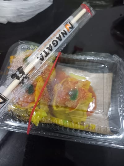 This media file shows a close-up, overhead view of a plastic container filled with what appears to be dim sum. The dim sum pieces are varied in color, with some being orange, yellow, and green. They are arranged in a dish, with condensation visible on the inside of the plastic lid.

A pair of chopsticks, still in their plastic wrapping, lies across the container. The wrapping is white with orange and black lettering. The words "NAGATA Bamboo Chopstick" are clearly visible, along with a small illustration of a panda. A red rubber band is stretched across the container, holding it closed.

The background is dark and out of focus, suggesting the photo was taken indoors or in low light conditions. The overall impression is of a takeout meal, possibly from a restaurant in Puloblang, Indonesia. There are no people visible in the image. The focus is entirely on the food and the accompanying chopsticks. The lighting is somewhat dim, and the image is slightly blurry, particularly around the edges.