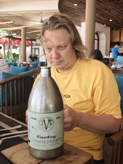 An adult Caucasian male with blonde hair and a light beard is seated at a table, looking down at a metal bottle in the foreground. He is wearing a yellow t-shirt with black lettering. The bottle is labeled "JVS Bartey UNFILTERED LAGER BEER". The background shows an outdoor dining area with wooden furniture and blue cushions, characteristic of a restaurant or cafe in Phan Rang – Tháp Chàm, Vietnam, as suggested by the presence of red lanterns and open-air architecture. Other patrons are visible in the distance.