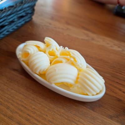 A dish of butter curls sits on a wooden table. The butter is in a white, elongated dish. The table has a wood grain pattern. A woven object is partially visible in the lower left corner of the image. The location is not evident from the image.