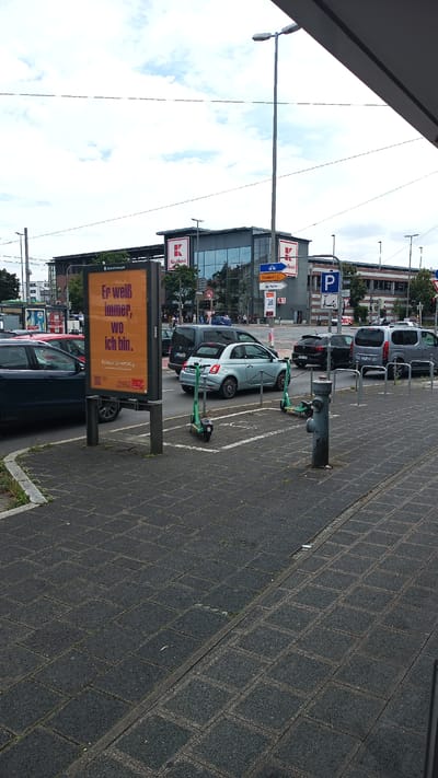 The image shows an outdoor scene with a street, sidewalk, and buildings in the background. In the foreground, there is a paved sidewalk with a planter box and a fire hydrant. There are several parked cars and two electric scooters on the sidewalk. In the background, there are buildings, including a Kaufland supermarket, and a street with traffic. There are also several streetlights and traffic signs visible. The sky is cloudy. The scene appears to be in an urban environment. Specifically, this appears to be the area near the Kaufland supermarket at Gustav-Adolf-Straße 150, 90425 Nuremberg, Germany, based on the billboard text and the signage. The billboard reads "Er weiß immer, wo ich bin." which translates to "He always knows where I am." Below this, it has a hashtag #DasIstGewaltig and a URL "stadtleben.me". The Kaufland signage is clearly visible.