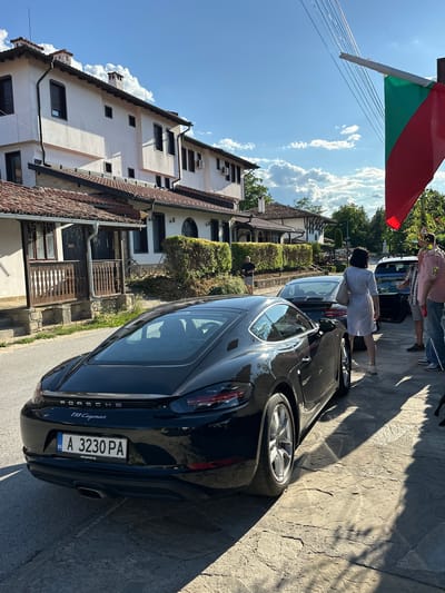 A black Porsche 718 Cayman is parked on a cobbled street in Arbanassi, Bulgaria. The car has a license plate that reads "BG A 3230 PA". In the background, traditional Bulgarian houses with tiled roofs and white walls are visible. A Bulgarian flag is partially visible on the right side of the image, hanging from a pole. Several people are standing near the car, some of whom appear to be talking or interacting with each other. The sun is shining, casting shadows on the street. The environment appears to be a historic village or town.