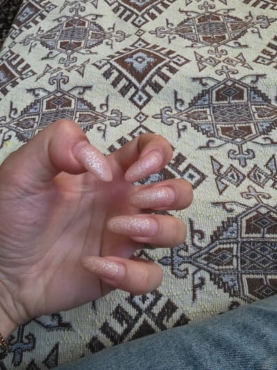 An overhead close-up depicts a left hand against a patterned textile background. The hand exhibits light to medium skin tone. All five visible fingernails are elongated, featuring a pointed almond or stiletto shape, and are coated with a shimmering champagne or light gold glitter polish, revealing natural nail growth at the cuticle line. A faint wristband is visible on the wrist. The background is a fabric adorned with a repeating geometric pattern in shades of brown, beige, and light blue-grey on an off-white base. A section of blue denim fabric is present in the lower right foreground. The scene is located in Staliyska mahala, Bulgaria.